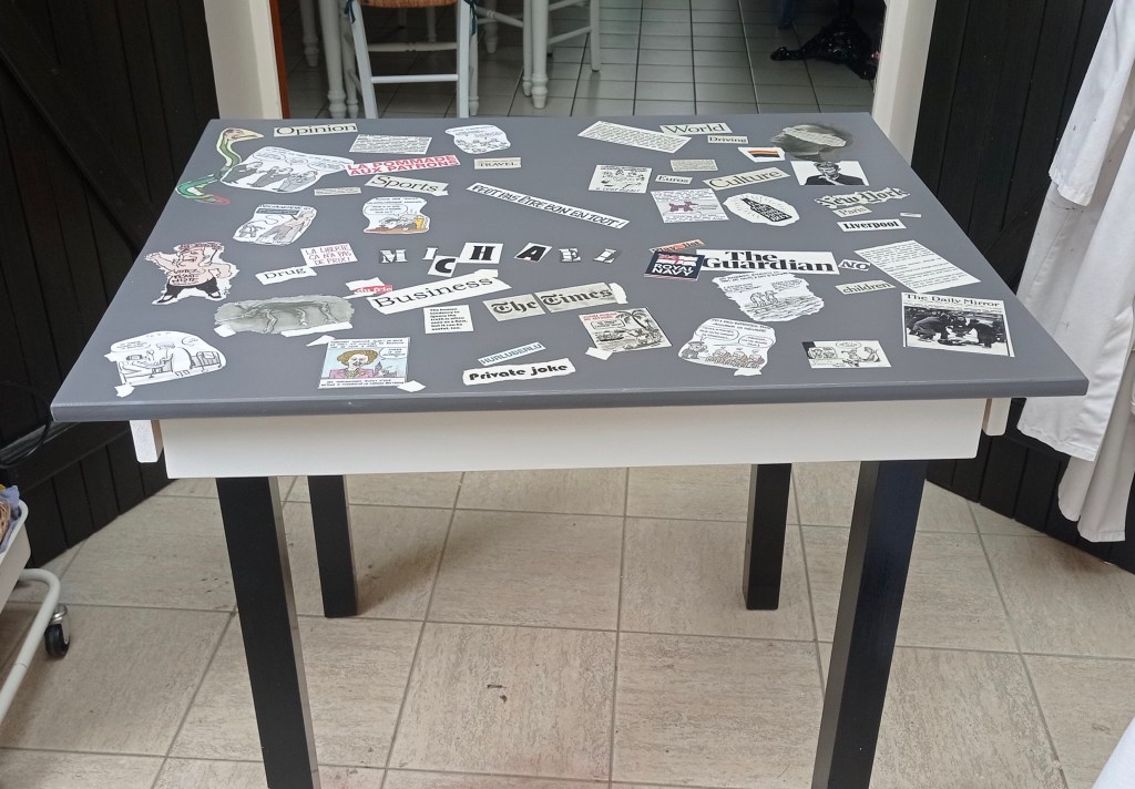 A table with a gray surface covered in various newspaper and magazine clippings, including headlines and illustrations, arranged in a random pattern.