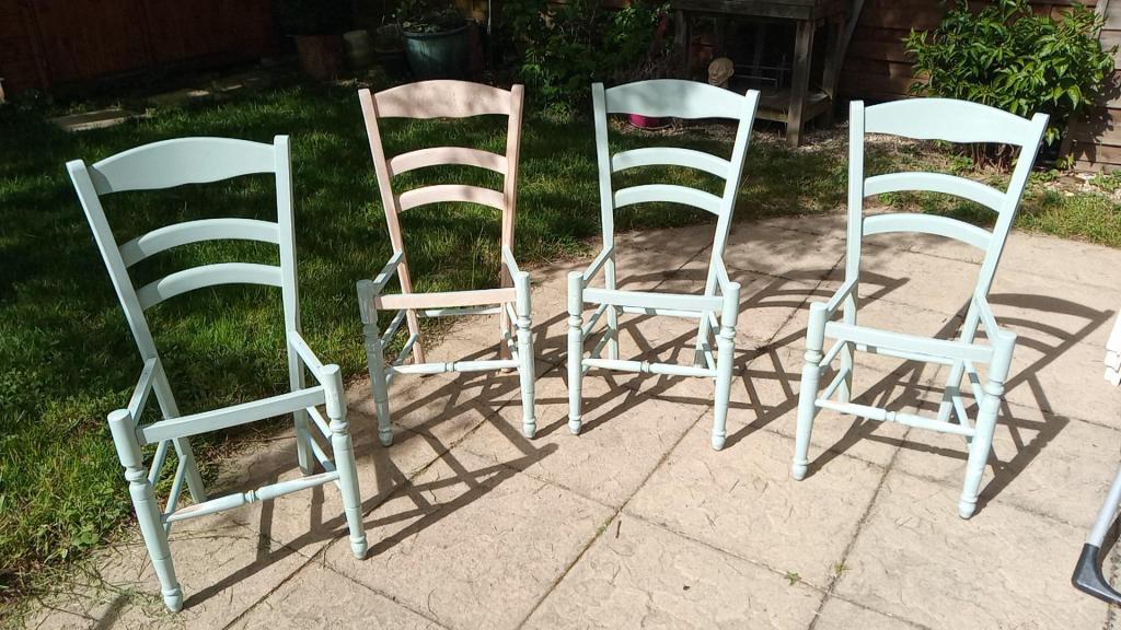 Four wooden chairs painted in pastel colors, arranged on a stone patio surrounded by grass.