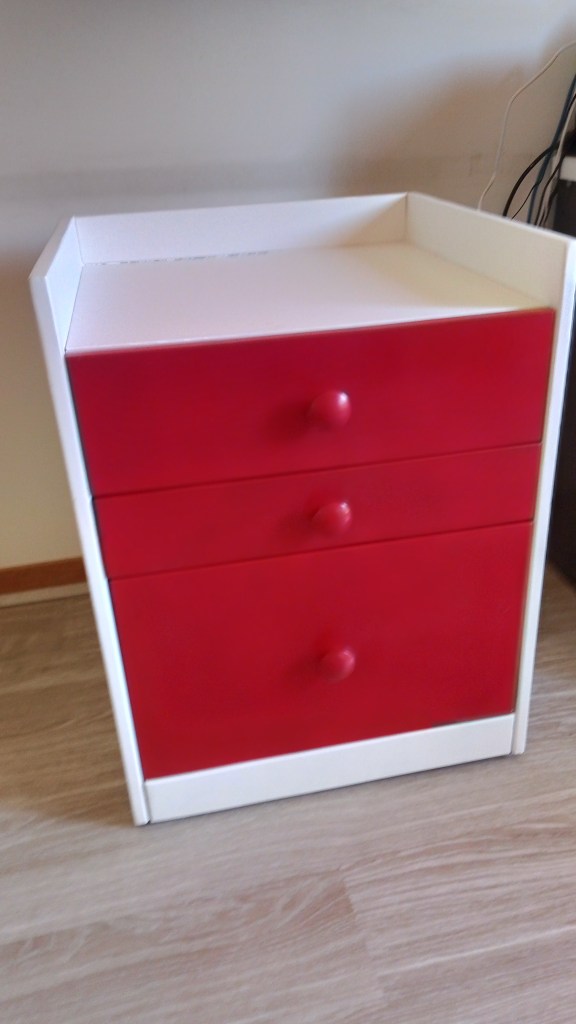 A three-drawer nightstand with a red front and white sides, placed on a wooden floor.