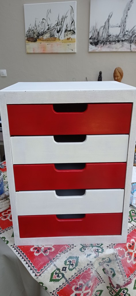 A compact storage box with four drawers, featuring a white exterior and alternating red and white drawer fronts, placed on a patterned tablecloth.