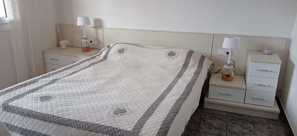 A neatly made bed with a light-colored quilt featuring a simple pattern, flanked by two nightstands with lamps in a minimalist bedroom.