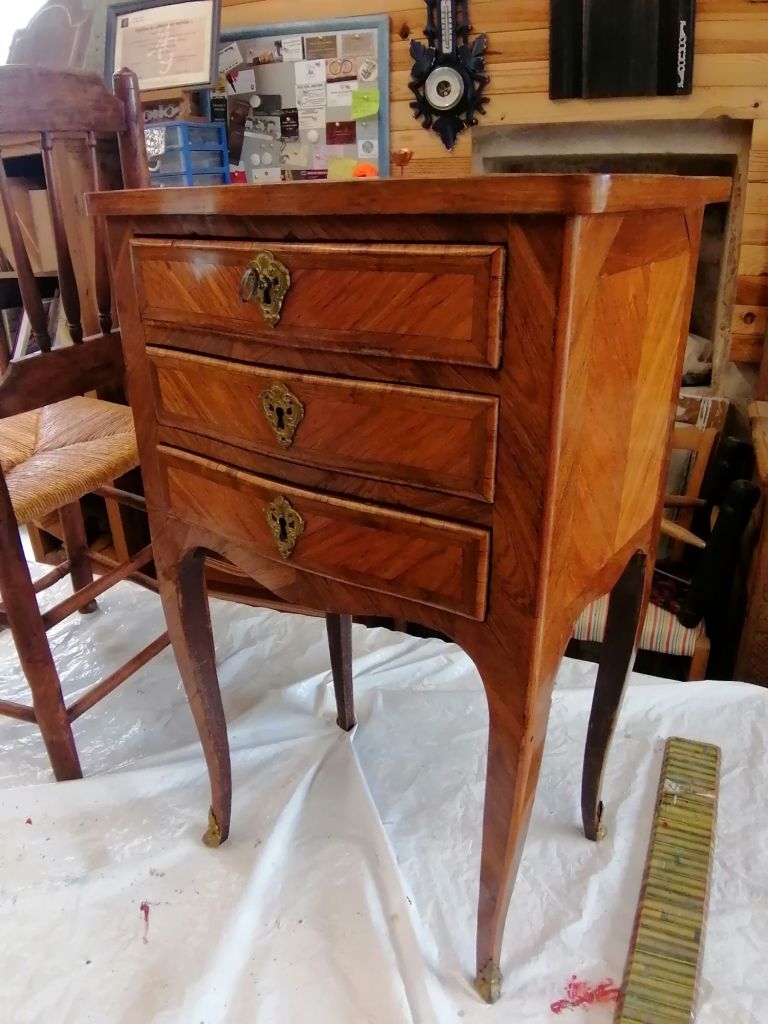 A vintage wooden nightstand with three drawers, featuring intricate inlaid patterns and ornate metal handles, placed on a white cloth background.