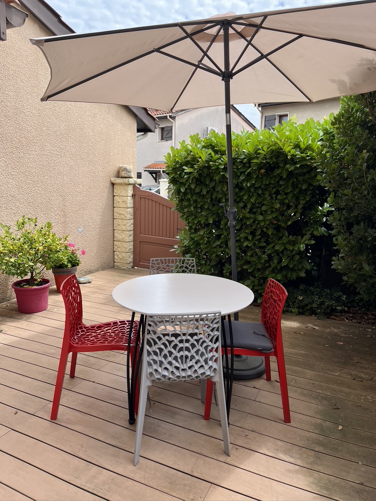 A patio scene featuring a round white table surrounded by four stylish chairs, two red and one gray, beneath a large umbrella. A green hedge and a doorway are visible in the background.