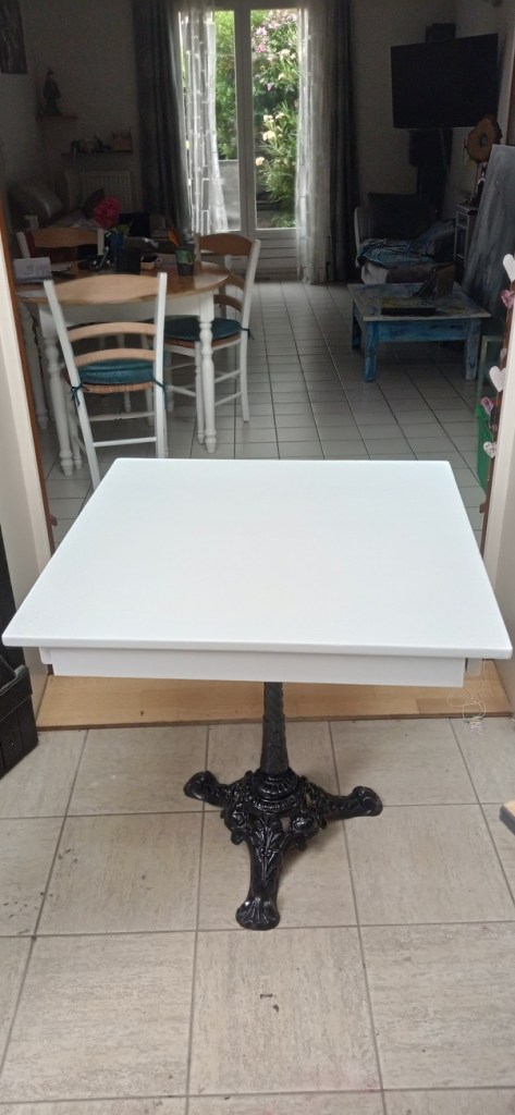 A white table with a decorative black base is positioned in a room with natural light coming through a window. In the background, there are chairs and a relaxed seating area.