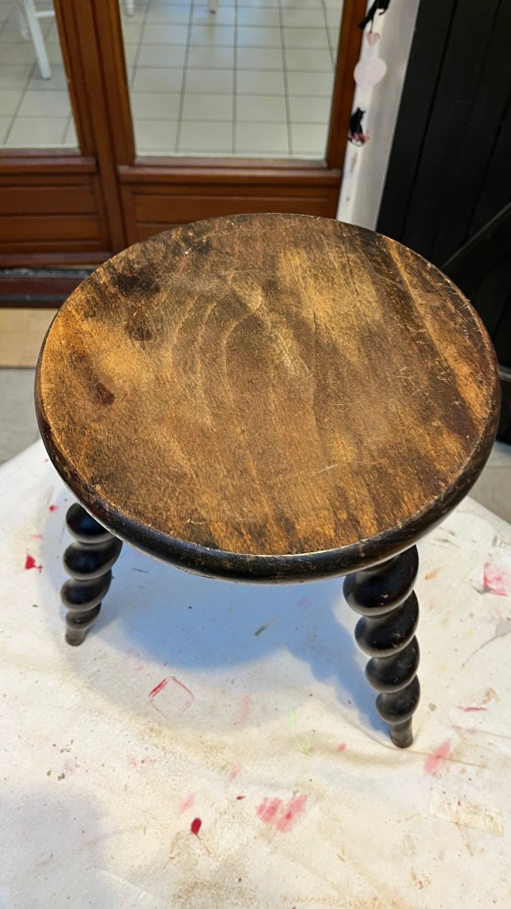 A wooden stool with a round top and spiral legs, set on a light surface, with blurred background showing part of a room.