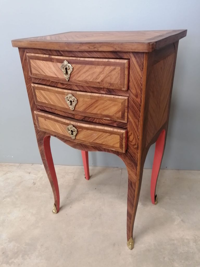 A vintage wooden nightstand with three drawers, featuring intricate geometric wood patterns and brass handles, sitting on pink legs.