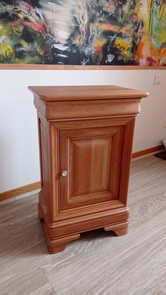 Wooden bedside table with a single door and a smooth top surface, placed against a wall with colorful artwork behind it.