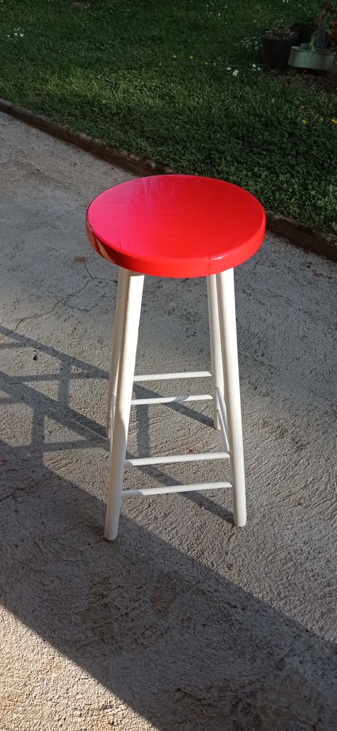 A red wooden stool with a round top and white legs set on a textured surface, surrounded by greenery.