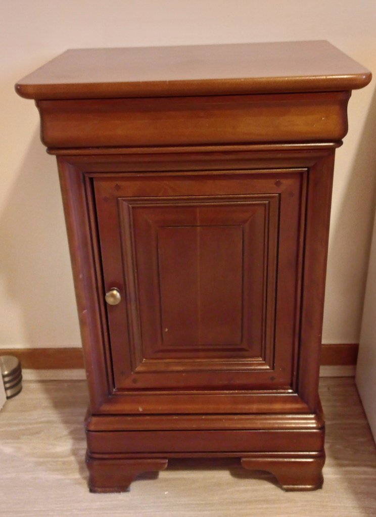 A wooden nightstand with a smooth top, a single cabinet door featuring decorative paneling, and a round knob.