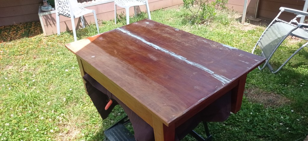 A wooden table outdoors with a partially finished surface, showing areas of wear and repair. Surrounding elements include grass and some outdoor furniture.