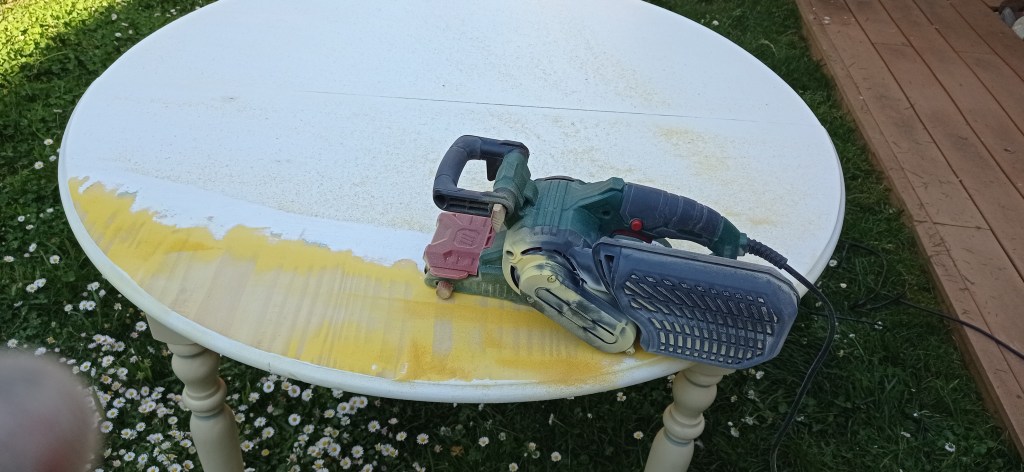 A circular white table in a grassy area with a sanding tool placed on top, partially sanded to reveal a yellow surface beneath. There are scattered white flowers around the table.