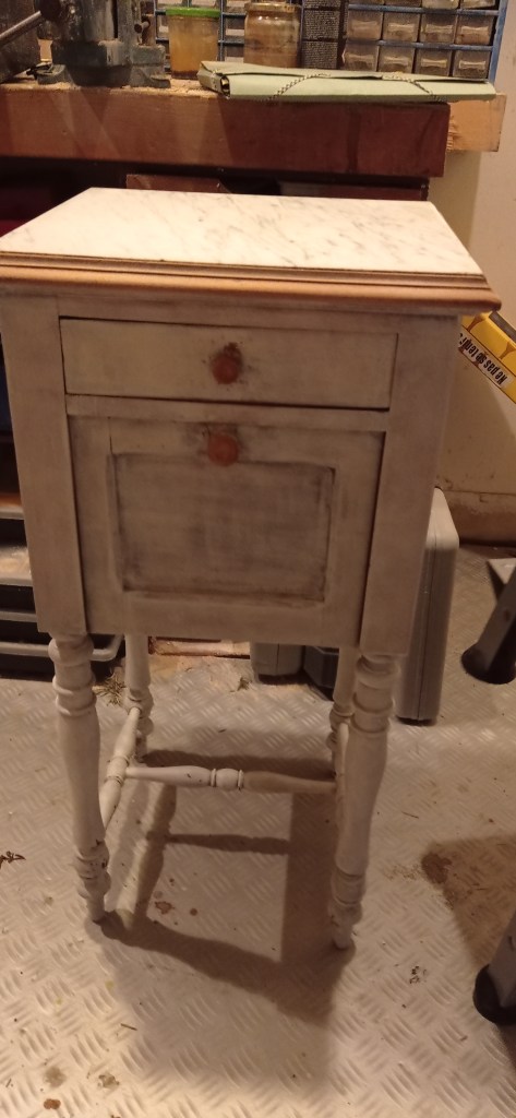 A small, vintage side table with a marble top, featuring a drawer and a cabinet below. The table has whitewashed wood and turned legs. It is placed in a workspace with tools visible in the background.