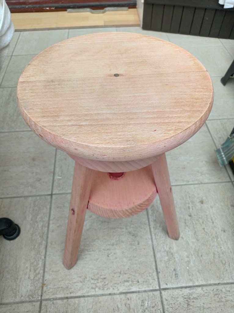 A wooden stool with a round top and three legs, featuring a pinkish hue and a simple design. The background shows a tiled floor and some furniture.