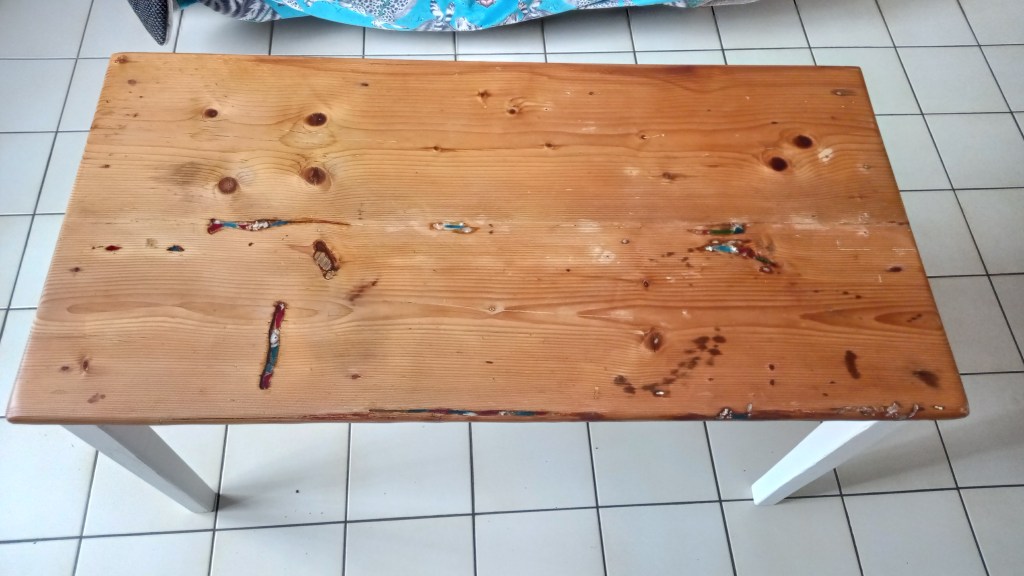 A wooden table with a rustic finish, featuring noticeable brown knots and colorful markings on the surface, set against a tiled floor.