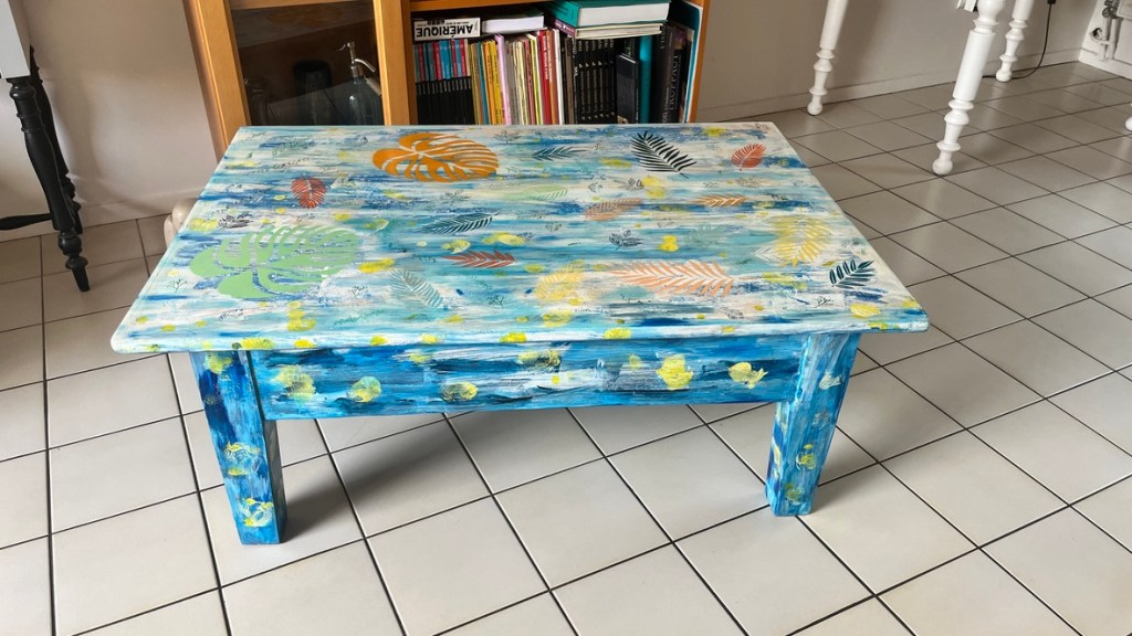 A colorful, hand-painted coffee table with a blue base and various leaf patterns in orange, green, and yellow on its surface, placed in a room with white tiled flooring.
