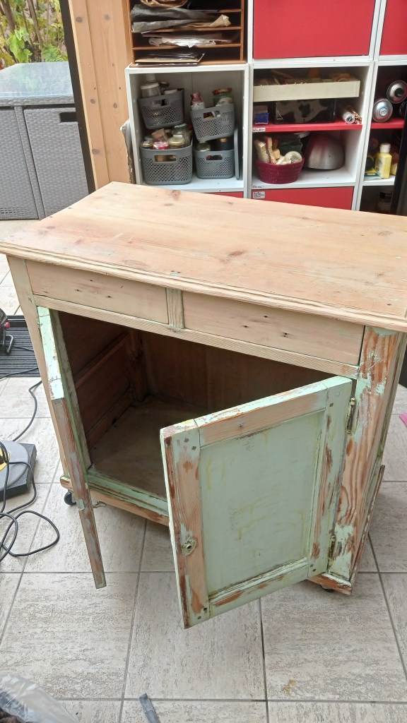 A wooden cabinet with one door open, revealing the interior. The cabinet has a rustic finish and is placed on a tiled floor, with storage bins and various items visible in the background.
