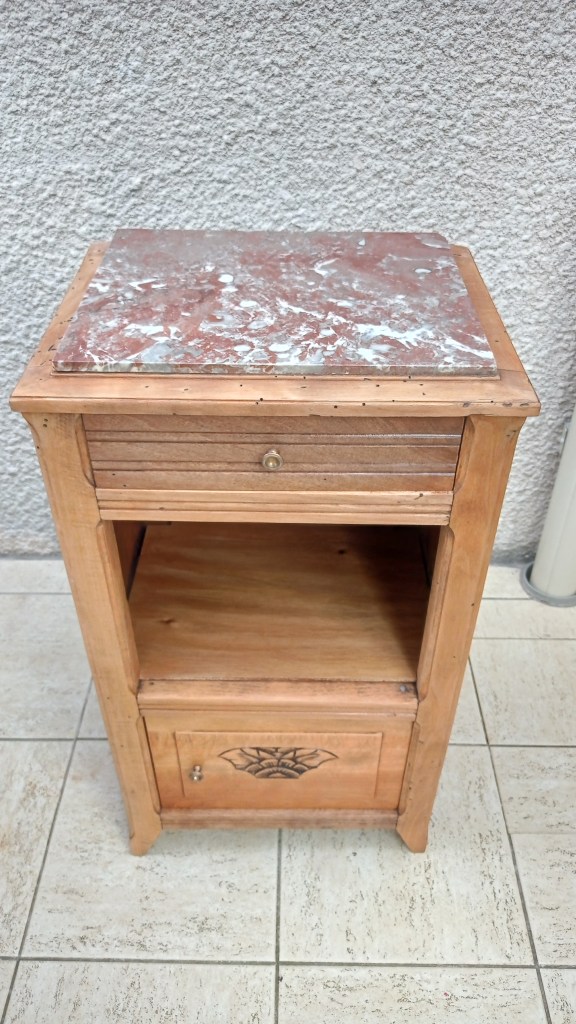 A wooden nightstand with a marble top, featuring a drawer and a lower compartment, positioned against a textured wall.