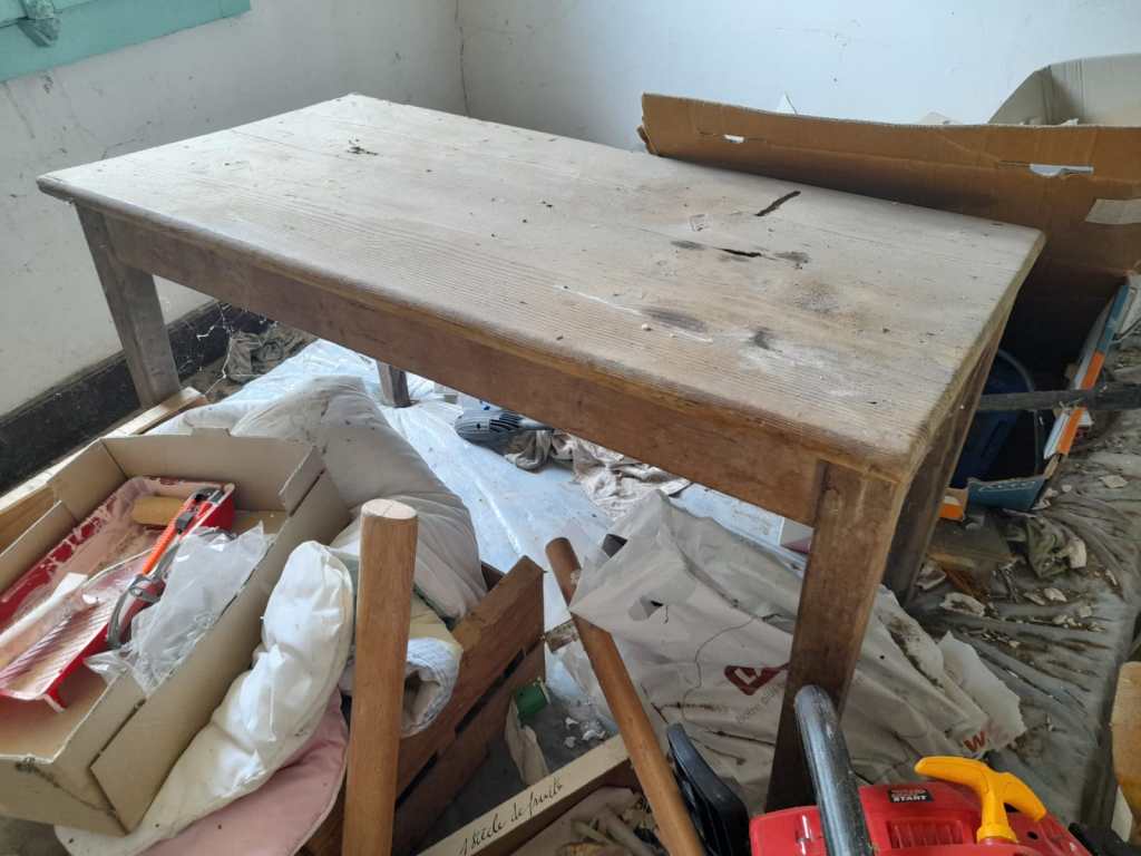 A worn wooden table placed amidst clutter, including tools and boxes, in a room with light-colored walls.