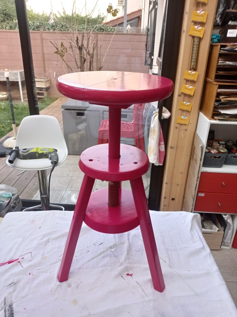 A pink wooden stool with a round top and three legs, featuring a central screw mechanism for height adjustment, placed on a white cloth with a background of greenery and outdoor furniture.