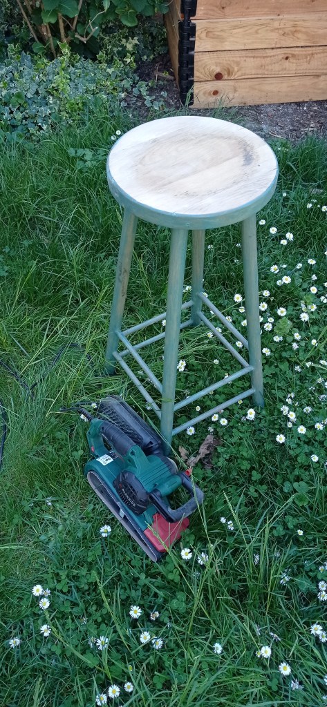 A green wooden stool placed on grass with small white daisies. A sander is lying on the ground next to the stool.