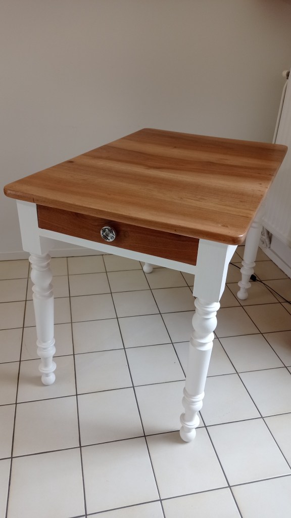 A wooden table with a light brown top and white legs, featuring a single drawer with a decorative handle.