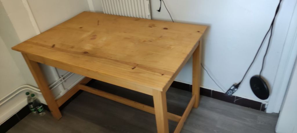 A simple wooden table with a smooth surface and four legs, placed against a wall, with a radiator and electrical outlets visible in the background.