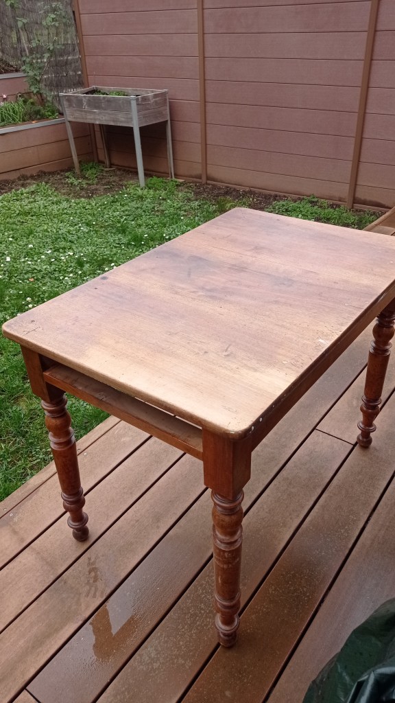A wooden table with a smooth top and turned legs, placed on a deck with green grass in the background.