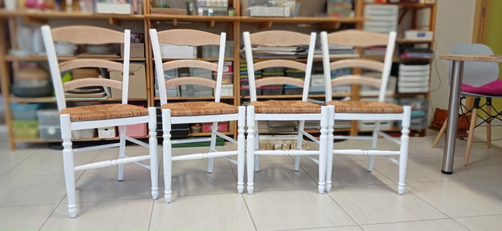 A set of three wooden chairs with white-painted legs and natural woven seats, arranged in a line against a backdrop of shelves filled with various items.