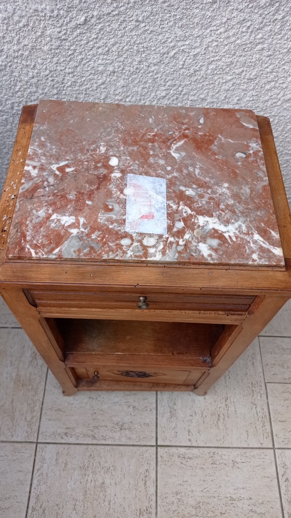A wooden nightstand with a red marble top, featuring a small drawer and an open shelf, placed on tiled flooring.
