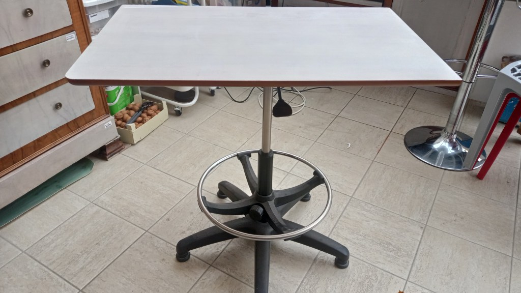 A simple, modern adjustable height table with a rectangular surface and a round base, set in a room with tiled flooring and nearby furniture.