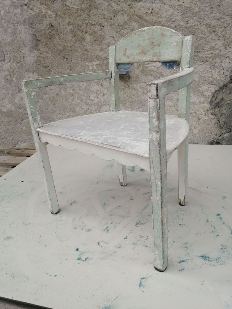 A vintage wooden chair with a chipped green and white paint finish, resting on a textured surface.