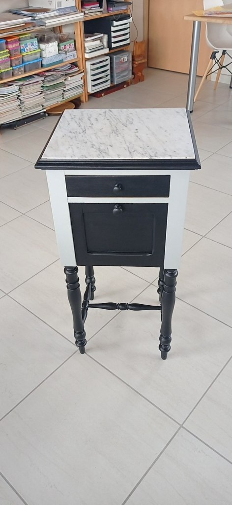 A small side table featuring a marble-patterned top, black painted legs, and a drawer with a knob. The background shows shelves filled with books and office supplies.