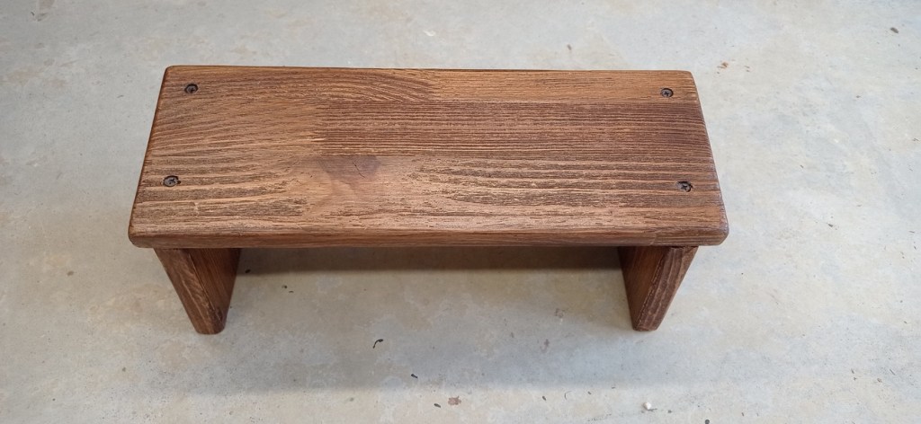 A simple wooden bench with a smooth top and sturdy legs, placed on a concrete floor.