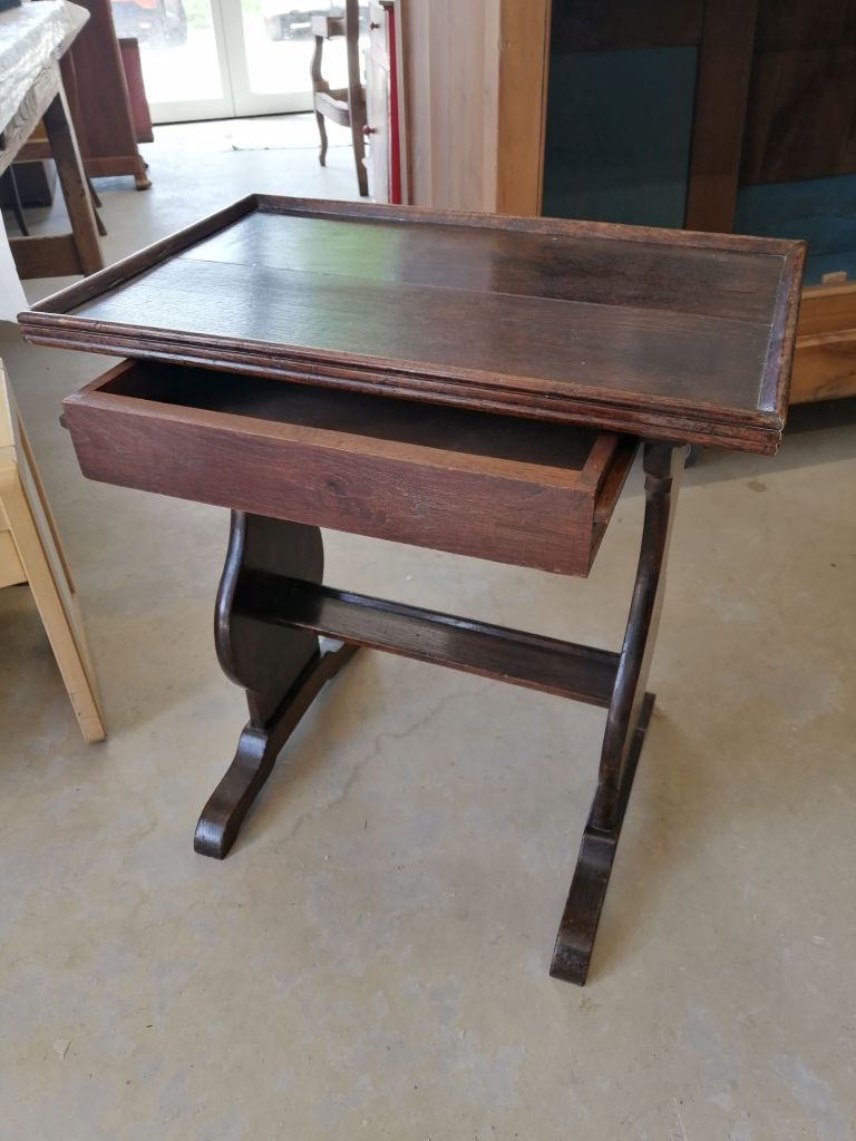 A wooden side table with a flat rectangular top and a single drawer, set against a backdrop of light-colored furniture and flooring.