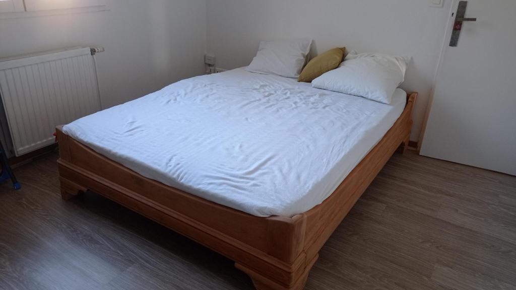A well-made wooden bed with white bedding and several pillows, placed in a minimalist room with light-colored walls and wooden flooring.