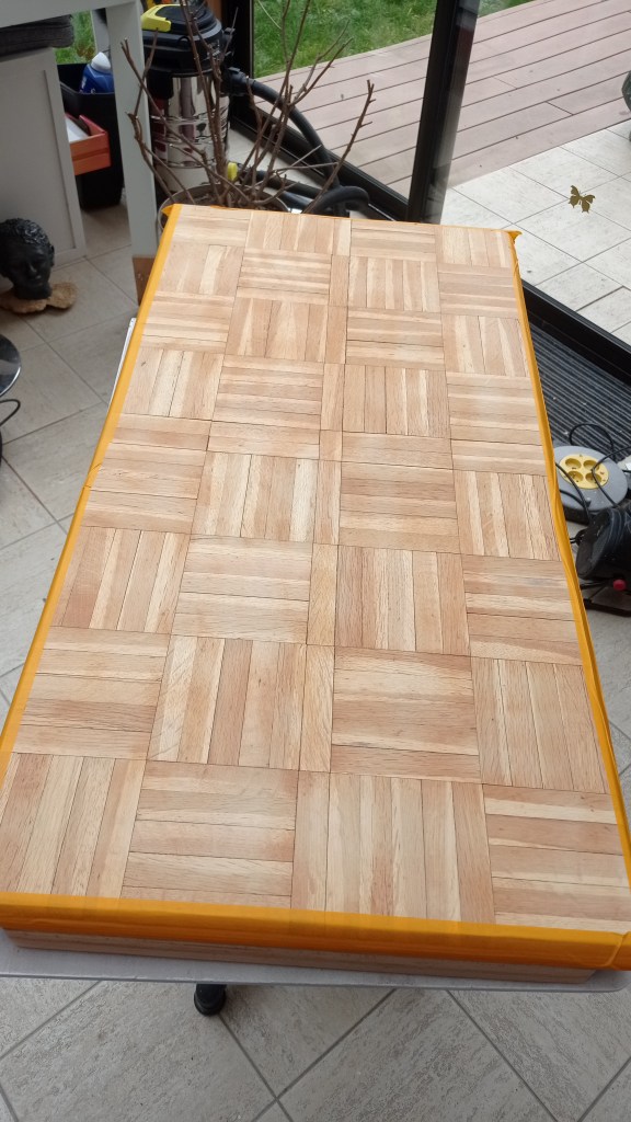 A wooden table with a parquet pattern on top, set against a background of a garden visible through a glass door.