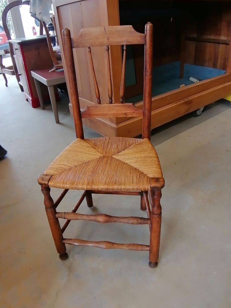 A vintage wooden chair with a woven straw seat, placed on a concrete floor surrounded by other furniture.