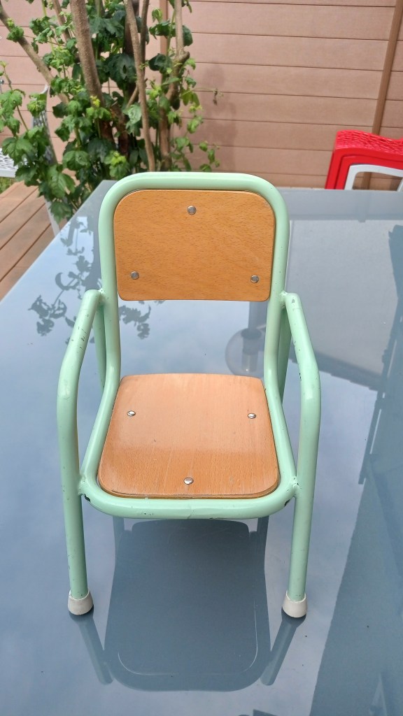 A small children's chair with a mint green metal frame and a wooden seat and backrest, placed on a glass table with a garden background.